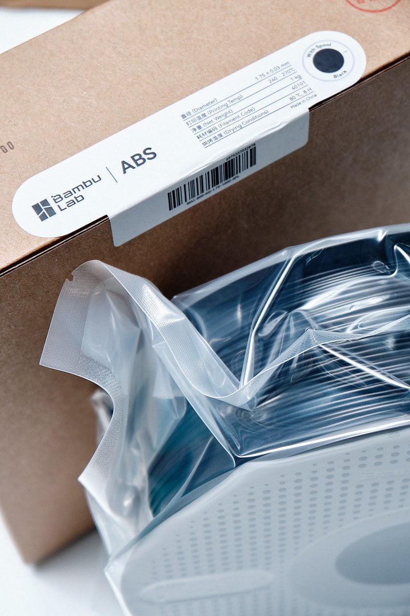Official Bambu Lab ABS Black filament packaging label and vacuum-sealed spool showing 1.75mm diameter, 240-270&deg;C printing temp, and 1kg weight.