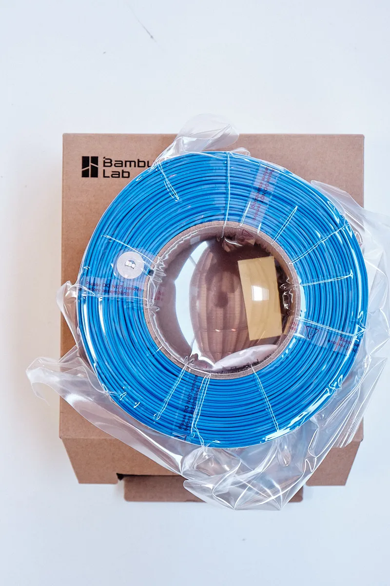 Top view of Bambu Lab PLA Lite Blue 1.75mm 1kg filament refill in vacuum-sealed packaging on its cardboard box.