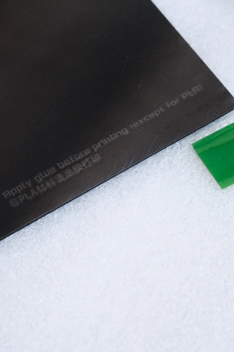 45-degree close-up of the lower-right corner of a Bambu smooth PEI build plate, shown on white foam packaging, with printed text “Apply glue before printing except for PLA” and a green marker indicating the peel-off protective film