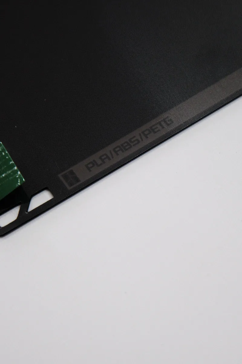 Close-up of the bottom area of a Bambu smooth PEI build plate showing printed filament compatibility markings for PLA, ABS, and PETG