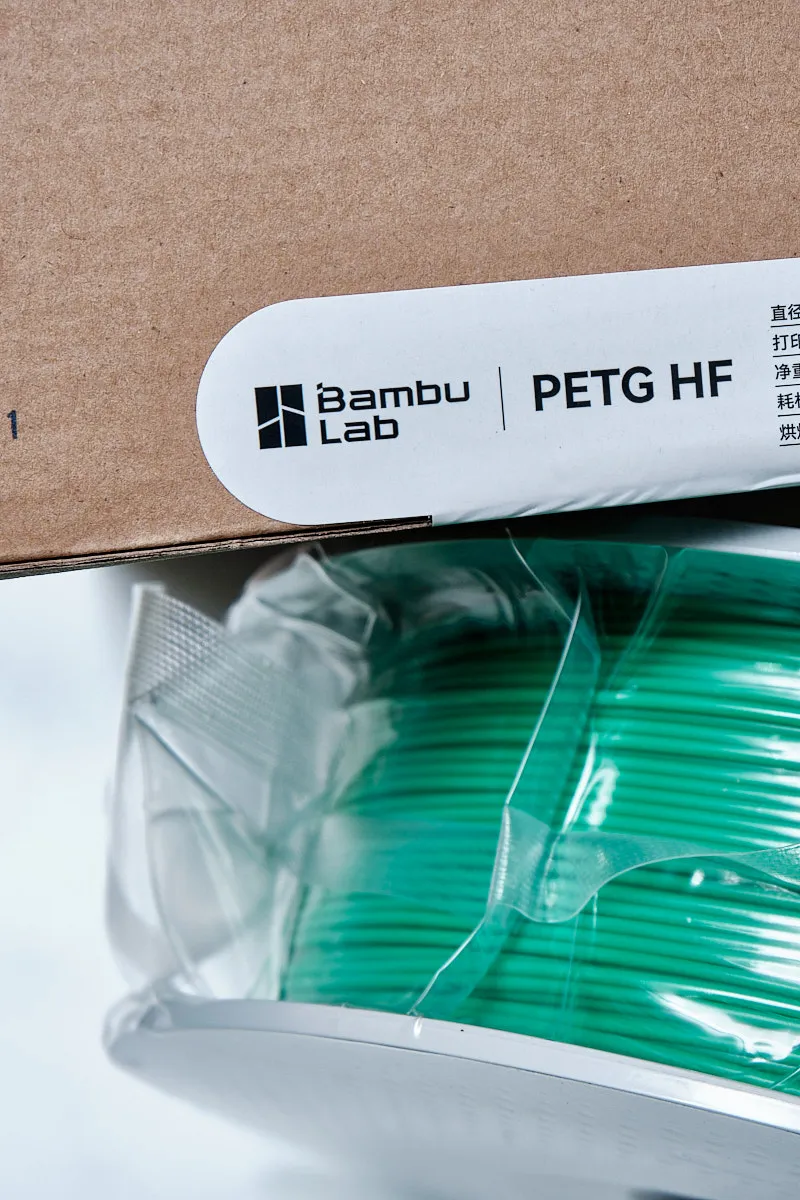 Close-up of Bambu Lab PETG-HF retail box label next to the vacuum-sealed green filament spool.