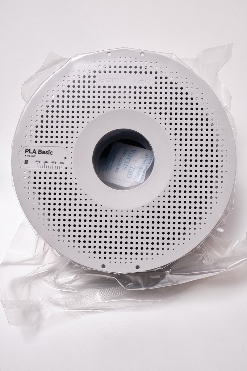 A top-down view of a white perforated Bambu Lab spool containing black PLA Basic filament, vacuum-sealed with a visible silica gel desiccant packet in the center.