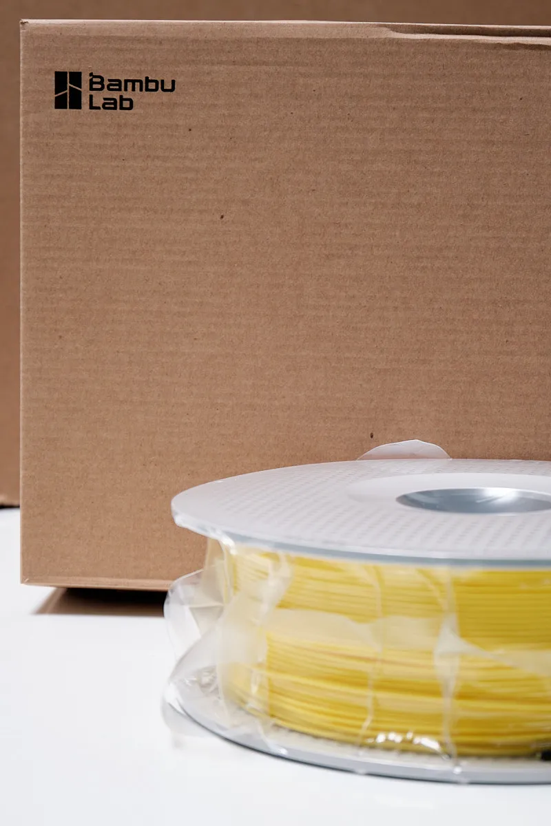 Bambu Lab PLA Basic Yellow filament spool in vacuum packaging next to its official brown cardboard shipping box with logo.