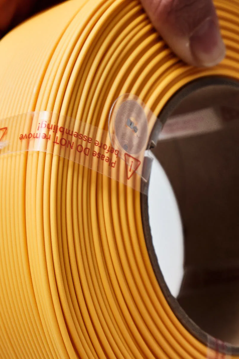 Macro shot of Bambu Lab PLA Lite Sunflower refill showing the integrated RFID intelligent identification chip and protective plastic assembly strap.