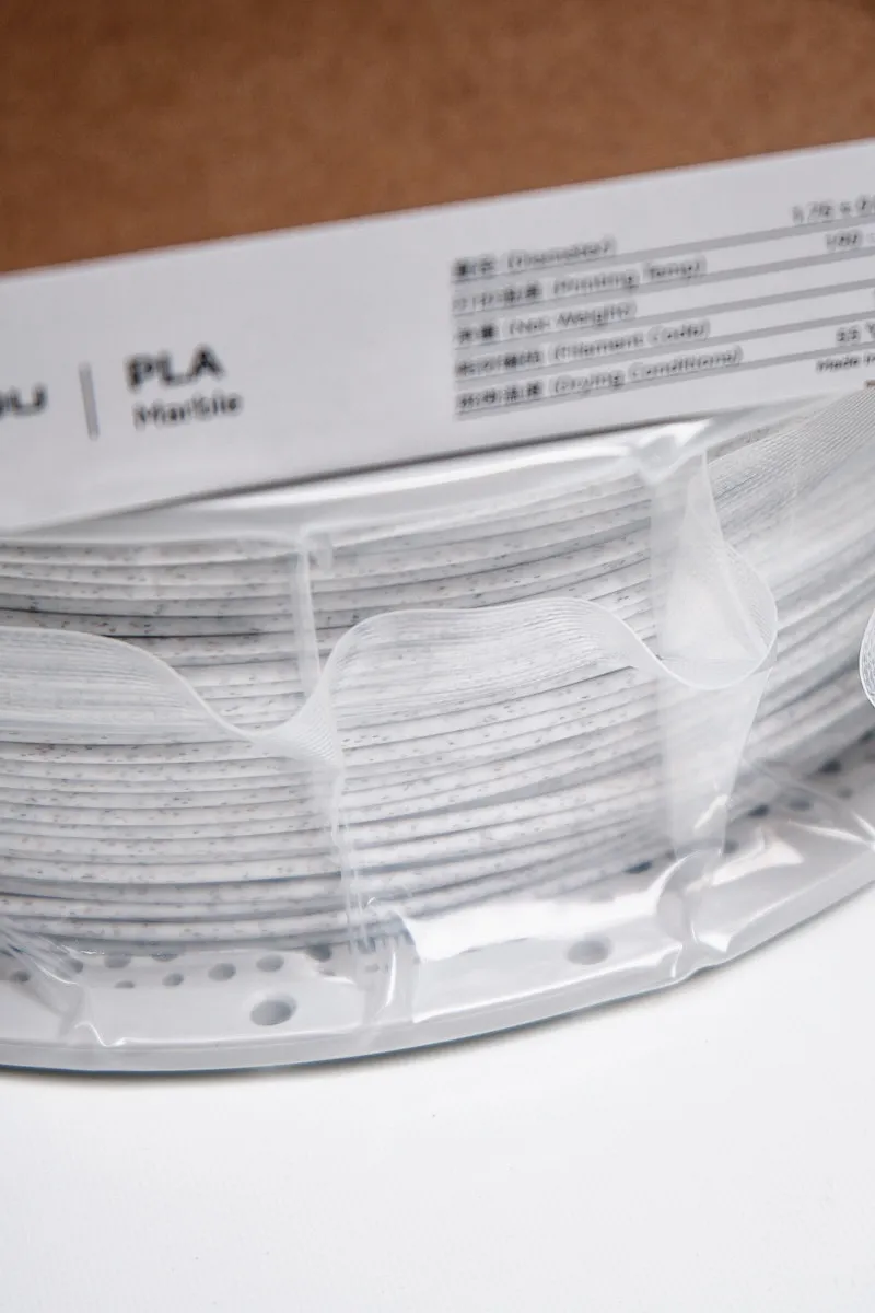 Close-up of White Marble PLA filament by Bambu Lab, showing the speckled stone-like texture and uniform winding through transparent vacuum packaging under a technical label.