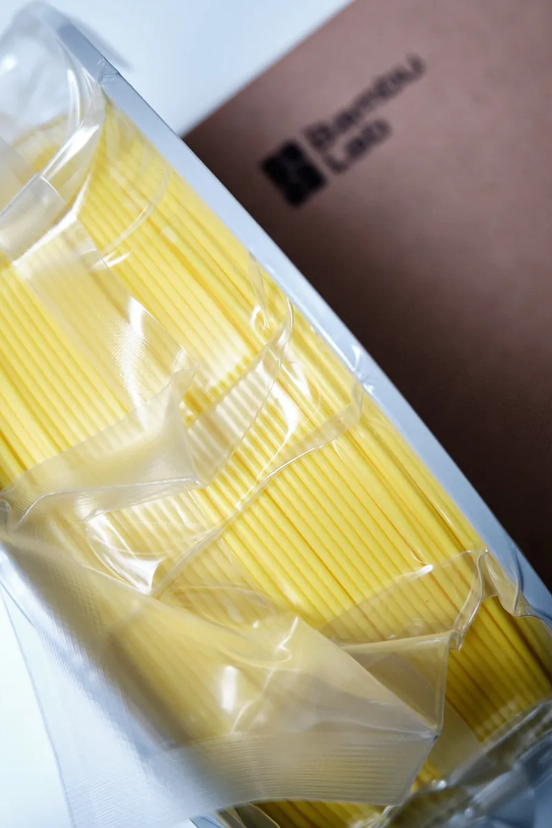 High-angle shot of vacuum-sealed Bambu Lab PLA Basic Yellow filament showing the protective plastic texture and branded box in the background.