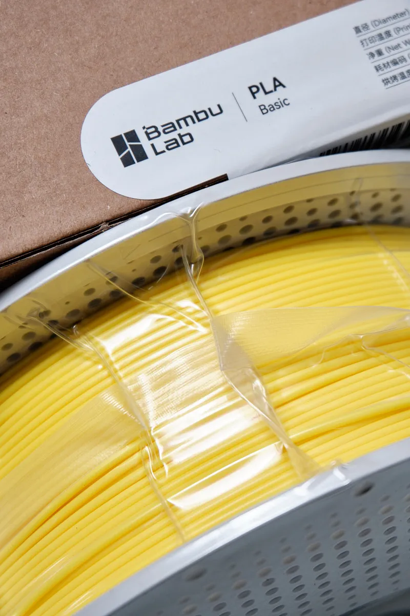 Close-up of Yellow PLA Basic filament spool with Bambu Lab logo, showing the white perforated spool and vacuum seal.