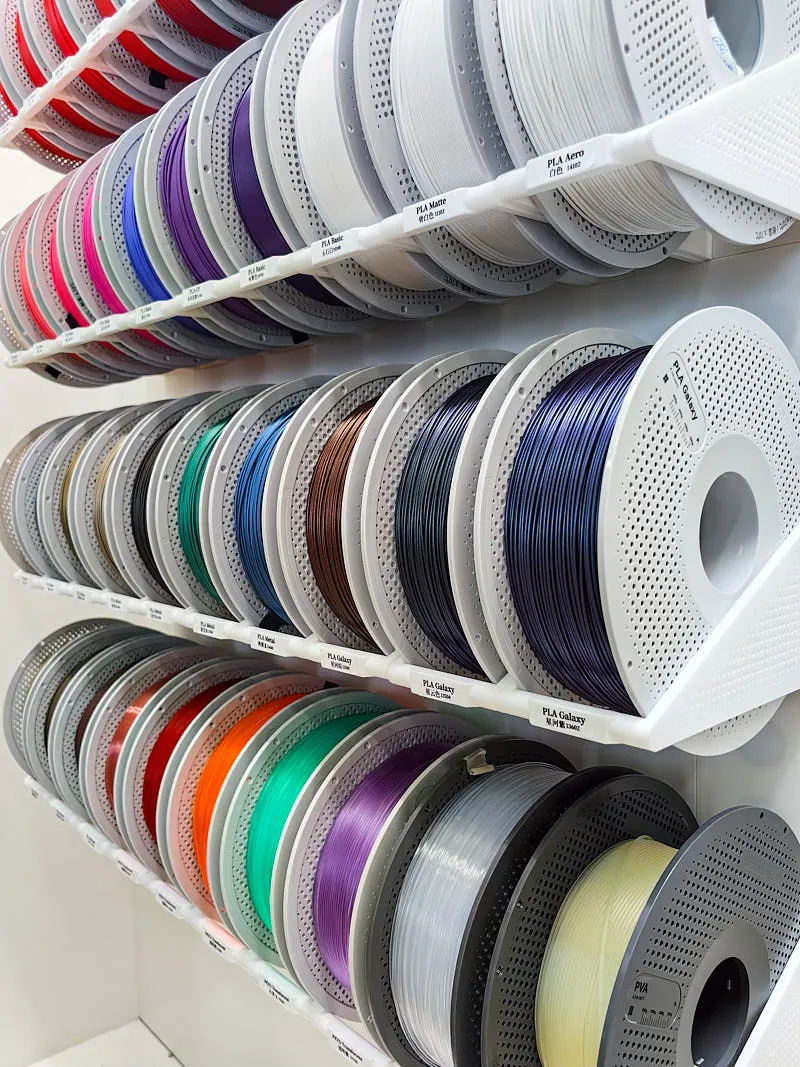 Rows of multi-color 3D printing filament spools organized on storage racks, showing a wide selection of PLA and specialty filaments
