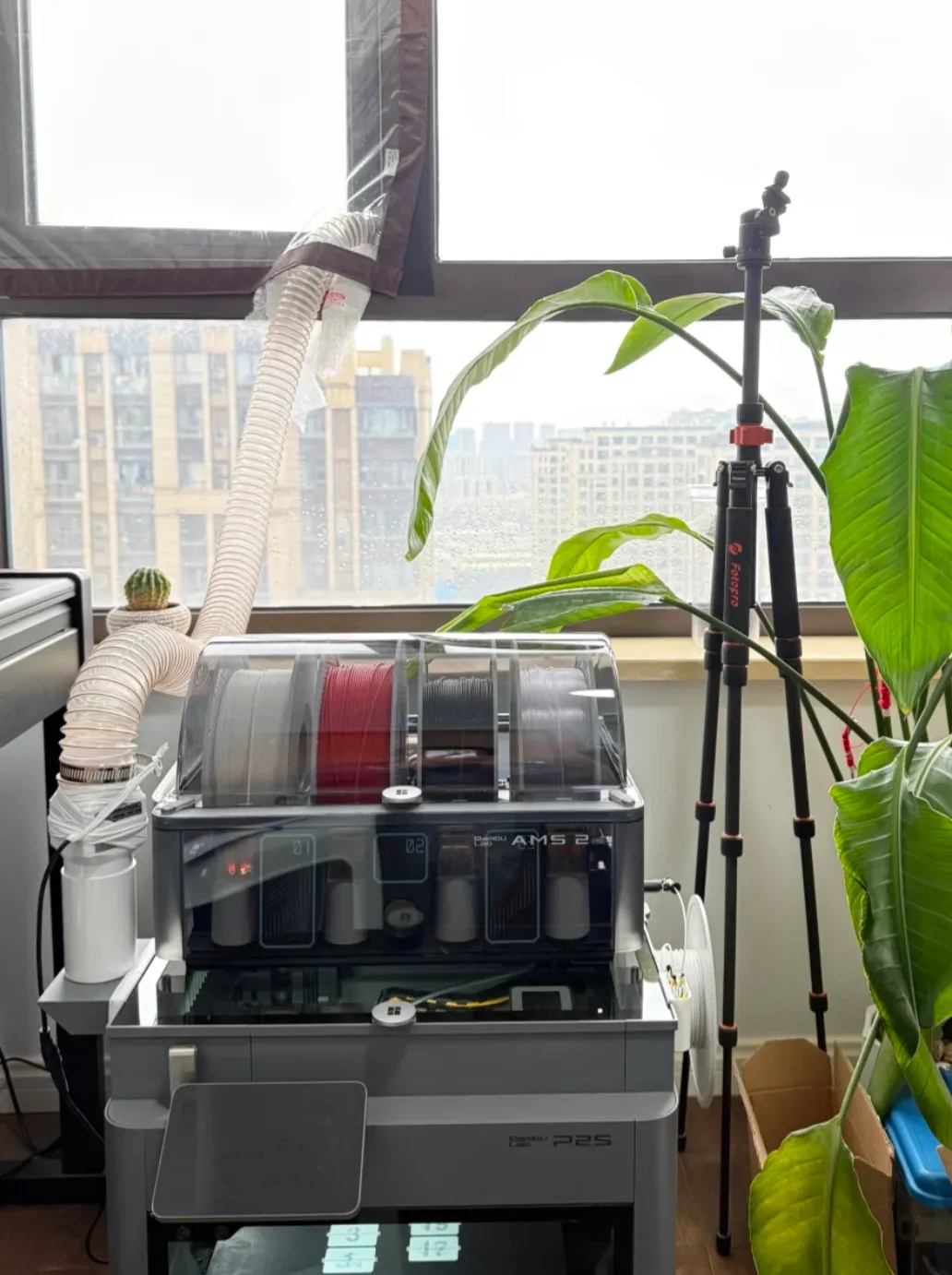 Bambu Lab P2S 3D printer placed near a window with exhaust duct venting outside